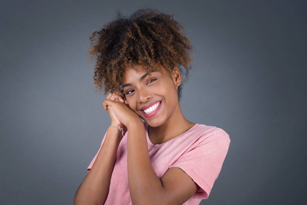 Dromerig Jonge mooie Afro-Amerikaanse vrouw met aangename uitdrukking, sluit de ogen, houdt handen gekruist in de buurt van gezicht, denkt aan iets aangenaams, poseert tegen grijze achtergrond. - Foto, afbeelding