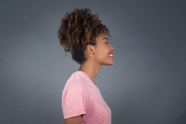 Perfil de mujer afroamericana sonriente con piel sana pura tiene expresión contemplativa, listo para tener paseo al aire libre, aislado sobre la pared del estudio gris con espacio de copia - Foto, Imagen