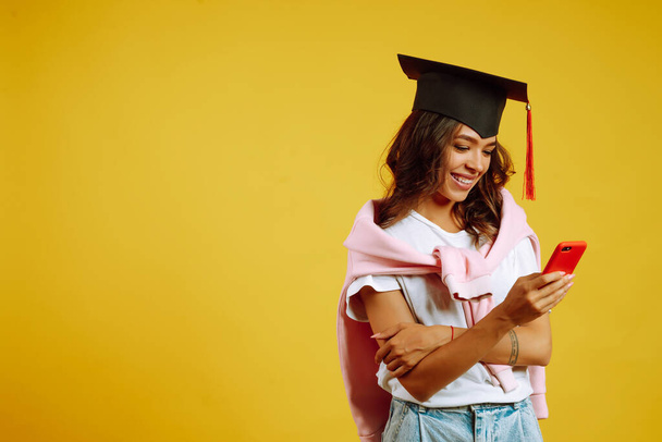 Graduate woman in a graduation hat posing with phone on a yellow background. African American woman with mobile  phone. Study, education, university, college, graduate concept. - Foto, immagini