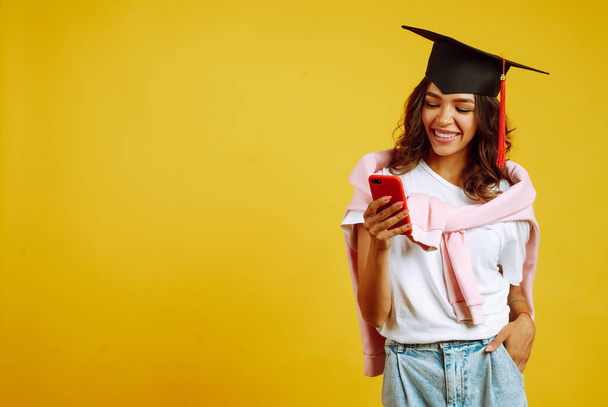 Mulher graduada em um chapéu de formatura posando com telefone em um fundo amarelo. Mulher afro-americana com telemóvel. Estudo, educação, universidade, faculdade, conceito de pós-graduação. - Foto, Imagem