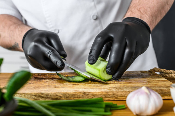 Egy fekete kesztyűs séf friss zöld uborkát szeletel egy fadaraboló asztalon. Az egészséges bioélelmiszerek főzésének koncepciója. - Fotó, kép