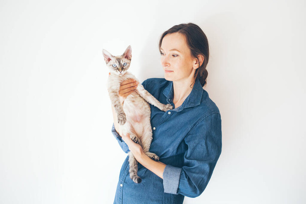 Styl życia zdjęcia ubranej kobiety w niebieskie dżinsy trzymającej ją i kota Devon Rex. Skupienie selektywne i naturalne światło - Zdjęcie, obraz