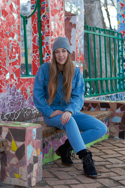 a woman in blue denim overalls and a gray hat sits on a bench near a wall of colored glass. - Photo, Image
