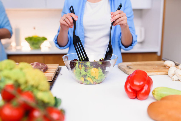Close up photo of young womans hands mixing fresh vegetables salad in a glass plate at the kitchen. - Valokuva, kuva