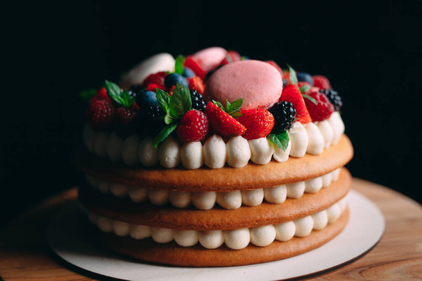 Fruit cake. Cake decorated with berries on a wooden stand on a black background. - Fotó, kép