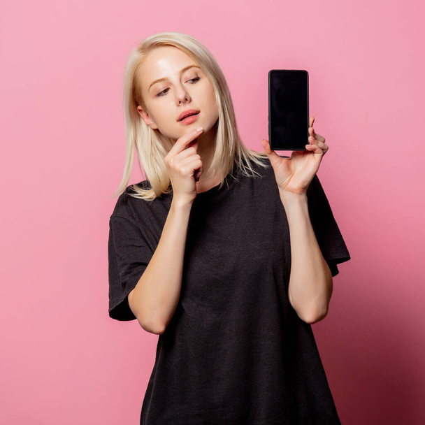 Femme blonde en noir moch-up tshirt et téléphone portable sur fond rose - Photo, image