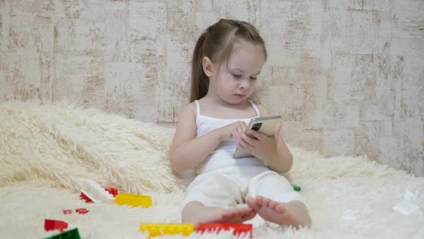 Lapsi leikkii puhelimessa istuessaan sohvalla suunnittelijan kanssa. Lelu älypuhelimessa. Elämä etäopetuksena esikoulussa. Moderni vauva - Materiaali, video