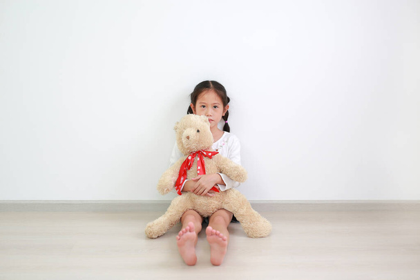 Peaceful asian little kid girl hugging a teddy bear doll sitting against white wall in the room alone. - Photo, image