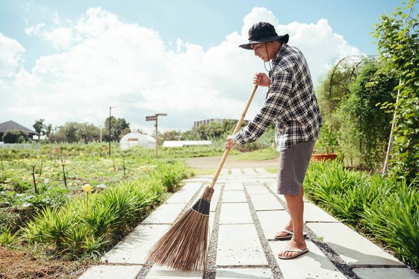 Asian elderly senior farmer cleaning garden with a broom stick in a farm. - Photo, Image