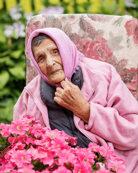 Photo of an elderly senior 85-year-old woman adjusts a pink kerchief on head among petunias and smiles sheepishly. Long-lived, retired, old age, wrinkles. Close up portrait - Photo, Image