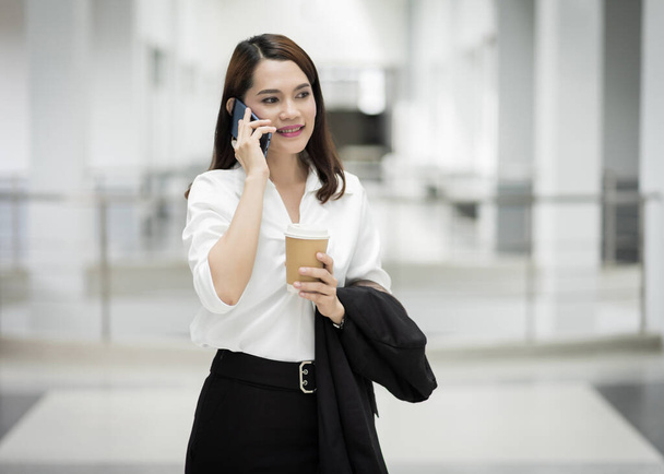 Retrato de una joven mujer de negocios asiática hablando por celular y sosteniendo una taza de café en un edificio de negocios. Foto de hermosa chica en suite casual con teléfono y taza de café. Foto de stock - Foto, imagen