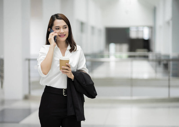 Portret młodej Azjatki rozmawiającej przez komórkę i trzymającej filiżankę kawy w budynku biznesu. Zdjęcie pięknej dziewczyny w casual suite z telefonem i filiżanką kawy. Zdjęcie z magazynu - Zdjęcie, obraz