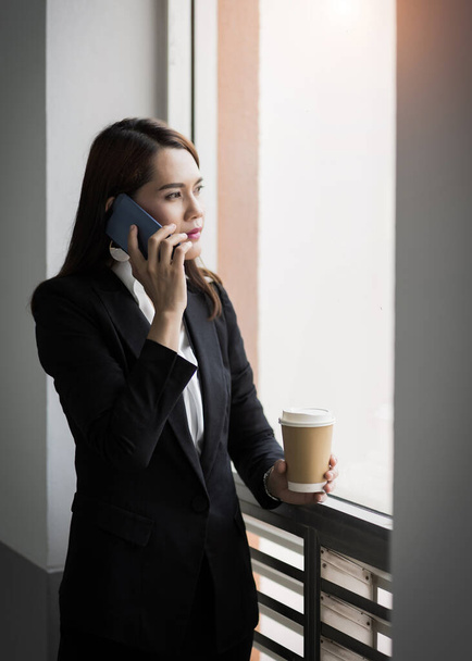 Portrait of a young Asian business woman talking over cellphone and holding cup of coffee in business building. Photo of beautiful girl in casual suite with phone and cup of coffee. Stock photo - Foto, imagen