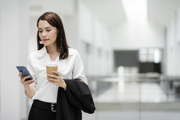 Portrait d'une femme d'affaires confiante en costume d'affaires tenant une tasse de café tout en utilisant un téléphone portable pendant la pause dans le bâtiment de l'entreprise. Photo de stock d'affaires. - Photo, image