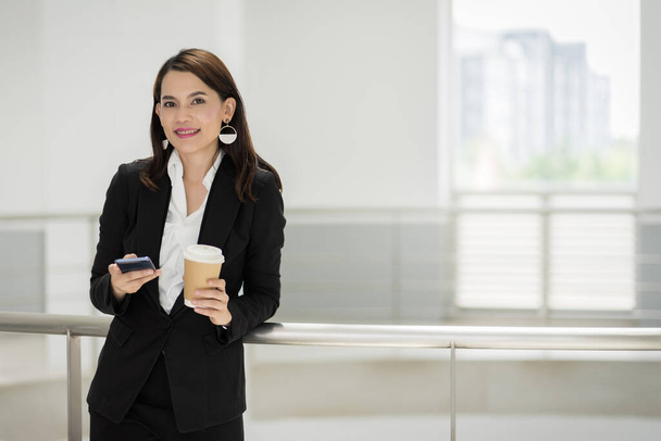 Portret van een zelfverzekerde zakenvrouw in een zakenpak die een kop koffie vasthoudt terwijl ze een mobieltje gebruikt tijdens de pauze in het bedrijfsgebouw. Bedrijfsvoorraadfoto. - Foto, afbeelding