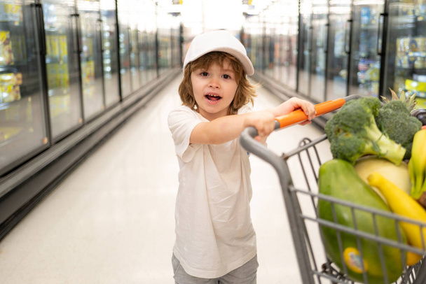 Joyful child boy in supermarket buys vegetables. Healthy food for children. Funny shopping. Cute kid with smile. Boy grocery shopping at the supermarket sitting in the cart helping his mother. - Photo, Image