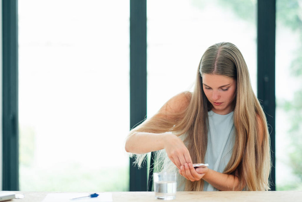 νεαρή λευκή γυναίκα κάθεται μπροστά από ένα ποτήρι νερό στο γραφείο εργασίας τραβώντας ένα χάπι από το περίβλημα - Φωτογραφία, εικόνα