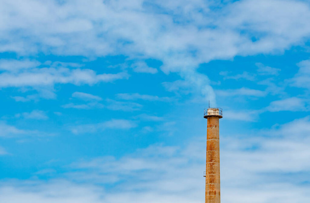 Ржавая дымовая труба завода против голубого неба и белых облаков. Белый дым из дымохода промышленных труб. Чистый воздух вокруг фабрики. Управление окружающей средой на заводе. - Фото, изображение