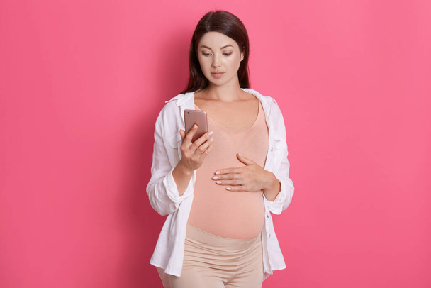 Pretty pregnant woman using her smart phone and touching her belly, looking at device's screen, looks concentrated, posing isolated over rosy background. - Fotoğraf, Görsel