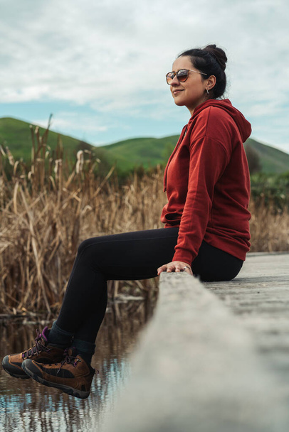 Nainen, jolla on rento vaatteet ja aurinkolasit istuu puinen kävelytie ja ympäröi keppi ja ryntää järvelle. Pystysuora kokovartalokuva - Valokuva, kuva