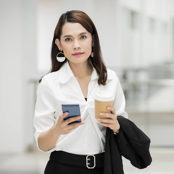 Portrait of a confident businesswoman in business suit holding a cup of coffee while using cellphone during break-time in the business building. Business stock photo. - Valokuva, kuva