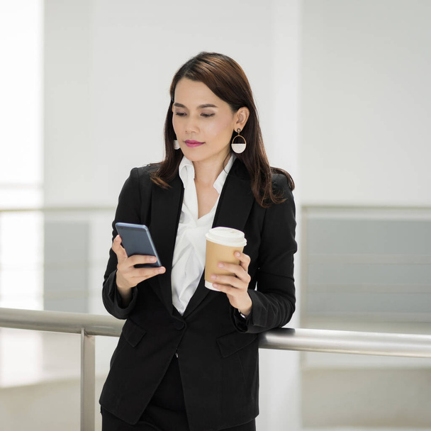 Porträt einer selbstbewussten Geschäftsfrau im Business-Anzug, die in der Pause im Geschäftshaus eine Tasse Kaffee hält und dabei das Handy benutzt. Archivbild aus der Wirtschaft. - Foto, Bild