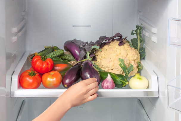 Woman takes the eggplant from the open refrigerator. woman's hand reaches for the vegetables on the refrigerator shelf. Weight loss diet. Healthy food. healthy lifestyle concept - Foto, afbeelding