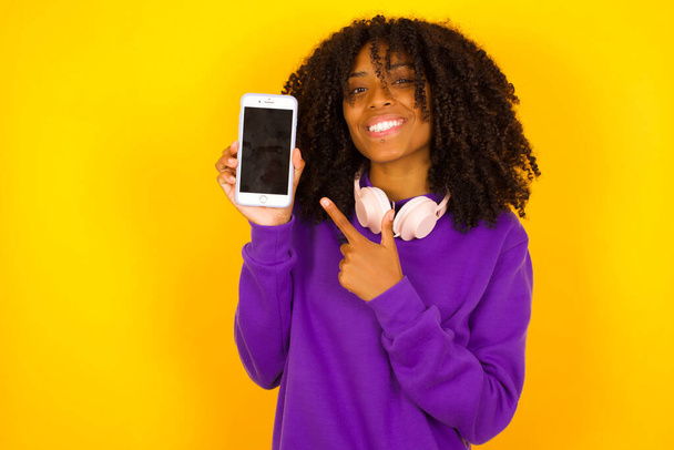 African american female  pointing on her mobile phone on yellow background. Emotions and gestures concept - Foto, Bild