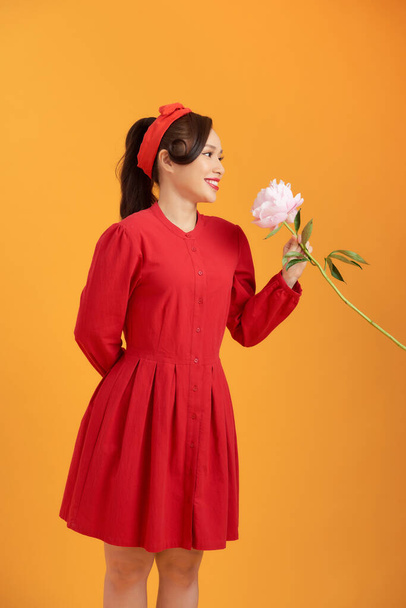 Encantador joven asiático en vestido rojo sosteniendo peonía flor aislada sobre fondo naranja. - Foto, imagen
