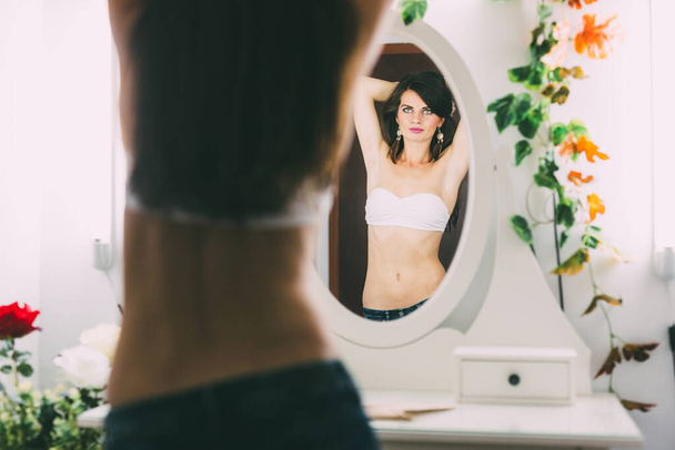 Beautiful female posing in front of the mirror, in a white bra and jeans, in a very seductive way - Foto, afbeelding