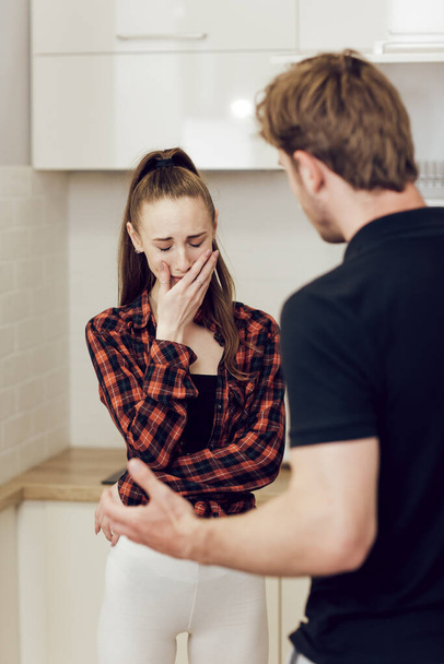 Problems of domestic violence and alcoholism in a young family. A drunk man shouts at his frightened wife at home in the kitchen and threatens her with his fist. A frightened young woman crying - Photo, Image