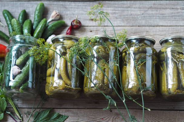 The benefits and harms of canned cucumbers. The process of home canning cucumbers in glass jars under lids for storage for the winter. Jars of cucumbers are in a row on a wooden background. - Фото, изображение