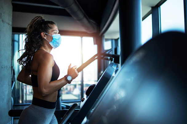 ジムでのトレッドミルでのスポーツ女性のトレーニングとcovid-19ウイルスの世界的なパンデミックの間にコロナウイルスから身を守るためにフェイスマスクを着用. - 写真・画像