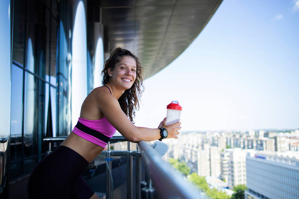 Smiling fitness woman with bottle of water resting on fitness center balcony after training. - Foto, afbeelding