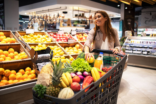 Hermosa mujer sonriente empujando el carrito de la compra y tomando frutas de los estantes en el supermercado. Comprar comida en la tienda de comestibles. - Foto, Imagen