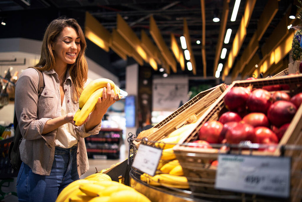 Woman enjoys buying healthy food in supermarket. Customer holding bananas at fruit department. - Photo, Image