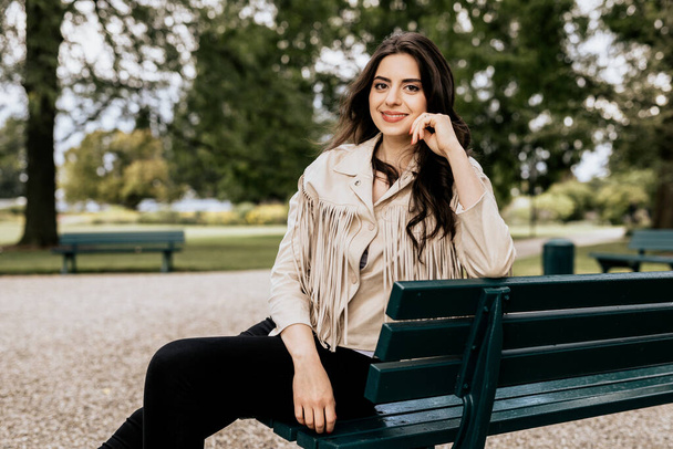 Beautiful attractive woman wearing beige fashion jacket sit outdoor. Young woman sit on the green bench with nature background - Foto, Imagem