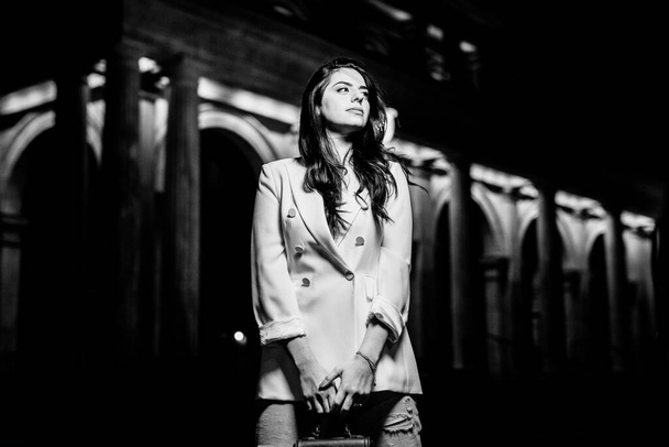 Donna attraente che indossa giacca di moda bianca e jeans distogliere lo sguardo. foto in bianco e nero di giovane donna su edificio con sfondo di illuminazione - Foto, immagini