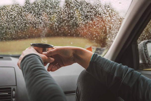 Close up shot of graceful female palms holding the hot tea tourist thermos mug. She sitting on co-driver seat inside modern car, enjoying the moody rainy day weather looking through windscreen drops. - Foto, imagen