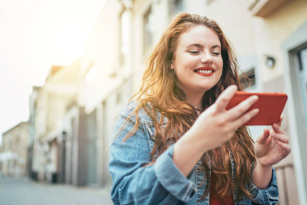 Smiling red curly long hair caucasian teen girl walking on the street and browsing the internet using the modern smartphone. Modern people with technology devices concept image. - Foto, afbeelding