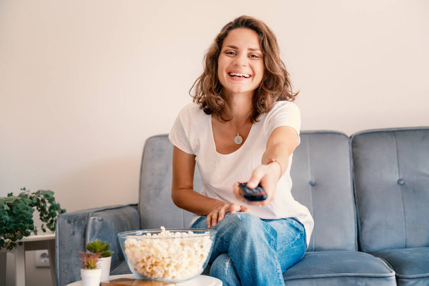Portrait of happy emotional beautiful young girl woman in jeans couch at home eating popcorn and watching TV in remote control in hands. - Valokuva, kuva