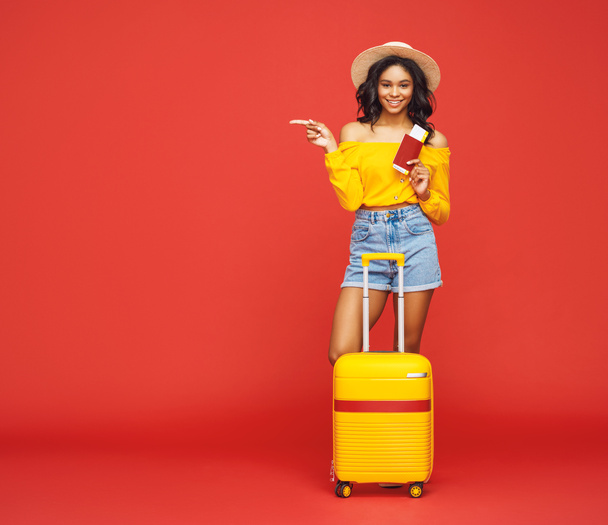Full body etnische vrouw met tickets en koffer glimlachend en wijzend weg tijdens de zomer reis tegen rode backdro - Foto, afbeelding