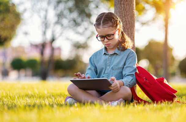 Side näkymä onnellinen tyttö lasit hymyillen ja lukeminen muistiinpanoja tabletin PC istuessaan nurmikolla lähellä puuta ja tehdä kotitehtäviä par - Valokuva, kuva