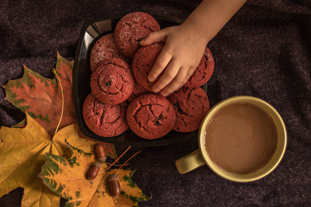 De hand van de kinderen reikt naar een bord cranberry koekjes. Stilleven met een kopje koffie, herfst multi-gekleurde esdoorn bladeren en eikels op de achtergrond van een paarse ruit. Herfstconcept. Bovenaanzicht. - Foto, afbeelding