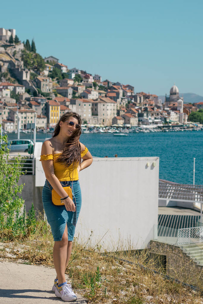 Attractive brunette wearing a blue skirt and an orange top posing outside near the sea on a warm summer day. Long hair flowing in the wind. Outline of Sibenik town in the distance. Influencer travels - Фото, изображение