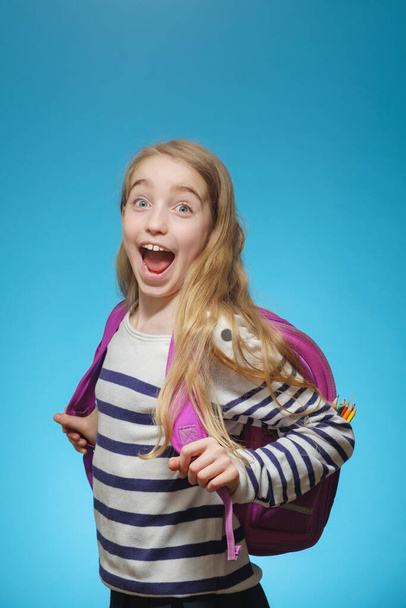Niña alegre caucásica con una mochila escolar sobre un fondo azul aislado. La chica está mirando a la cámara y sonriendo. - Foto, imagen