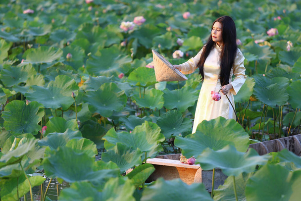 Vietnamesin in ao dai traditionelles vietnamesisches gelb-weißes Kleid im Lotusteich mit altem Boot aus Holz. - Foto, Bild