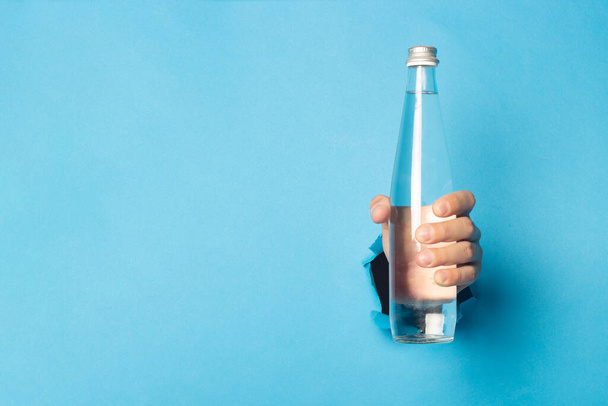 Mano del hombre sosteniendo una botella de vidrio con agua sobre un fondo azul brillante. - Foto, Imagen