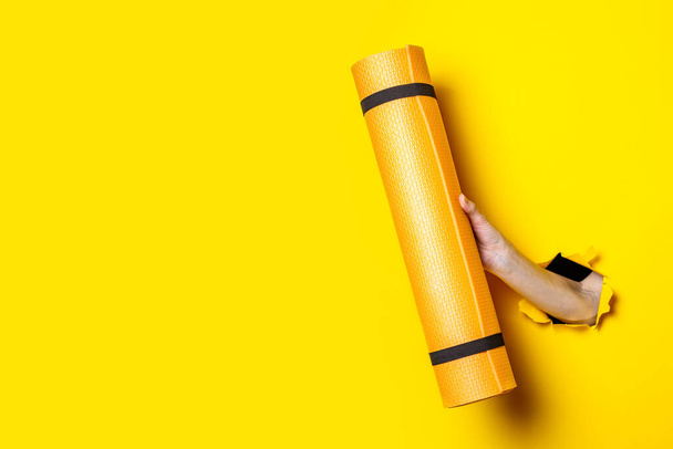 Mão feminina segurando um tapete de ioga laranja em um fundo amarelo brilhante - Foto, Imagem