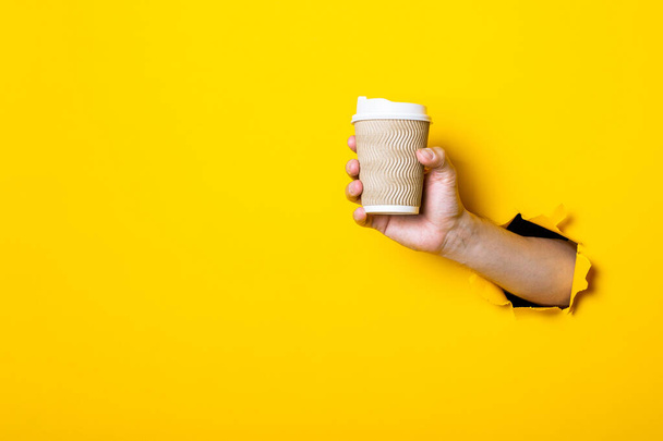 Το χέρι του ανθρώπου κρατά ένα χάρτινο κύπελλο με καφέ σε φωτεινό κίτρινο φόντο. - Φωτογραφία, εικόνα